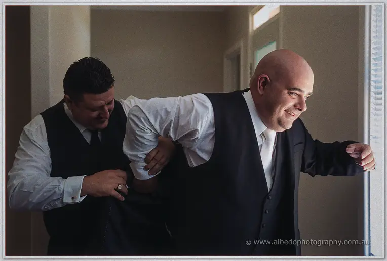 A groomsman slides James's suit jacket on as he laughs toward the window light