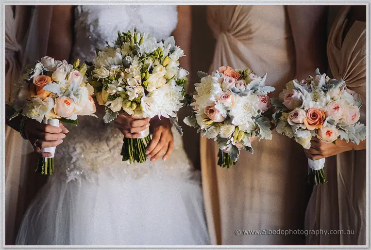 Elena and her bridesmaids hold peach-rose and white-freesia bouquets against their gowns