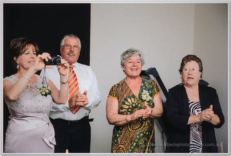 A woman raises a compact camera while three older relatives smile and clap beside her