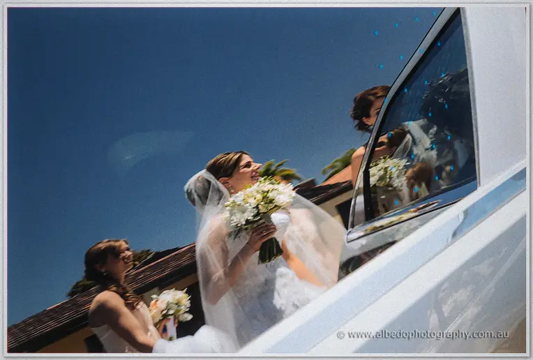 Veiled Elena laughs with her bouquet as bridesmaids step out of the white car under blue sky