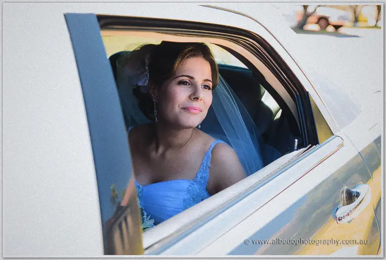 Veiled Elena in a pale blue gown smiles from the rear window of the cream bridal car