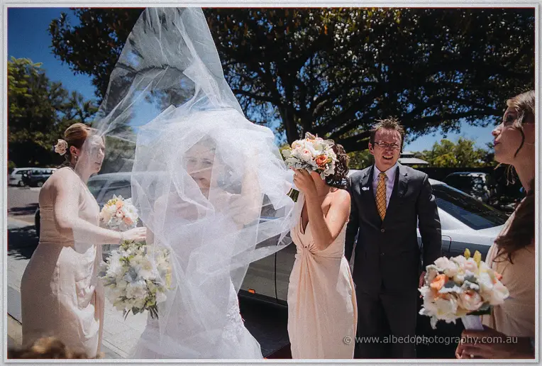 Wind whips Elena's veil over her face as bridesmaids and guests laugh beside the limousine