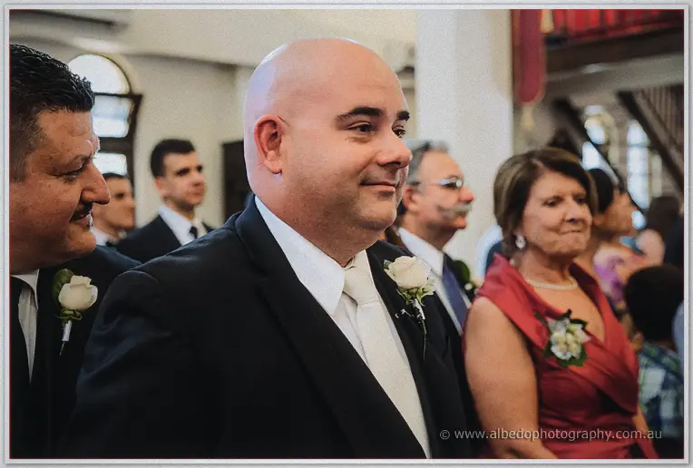 James smiles softly among standing guests, white rose boutonniere on his black suit