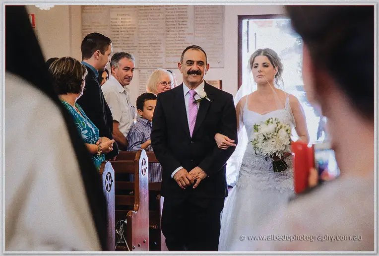 Elena's mustached father beams as he walks her down the aisle past risen guests