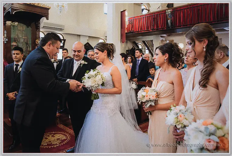 A groomsman places the ring on Elena's finger as James and blush bridesmaids watch