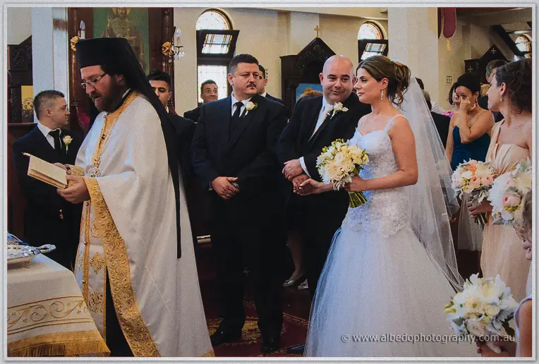 Elena and James hold hands before the white-and-gold vestmented priest as bridesmaids watch