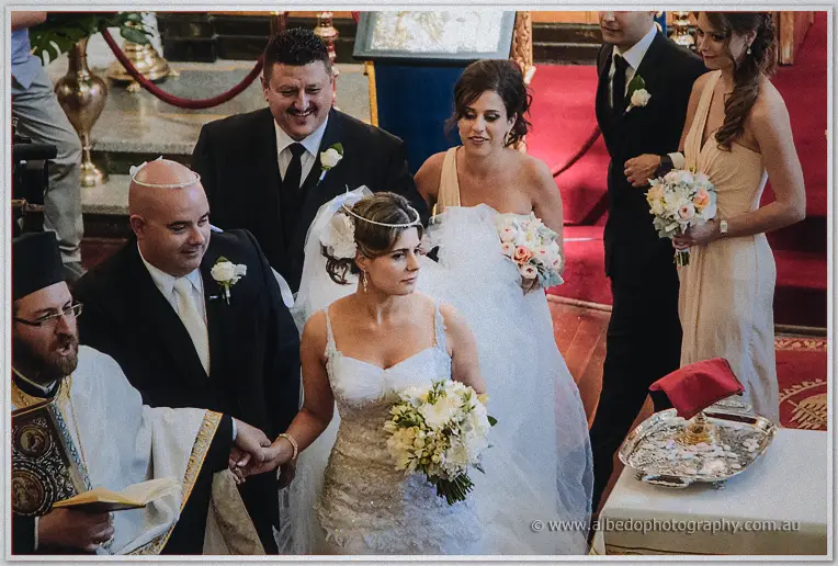 Crowned Elena leads James hand in hand around the ceremony table as guests watch