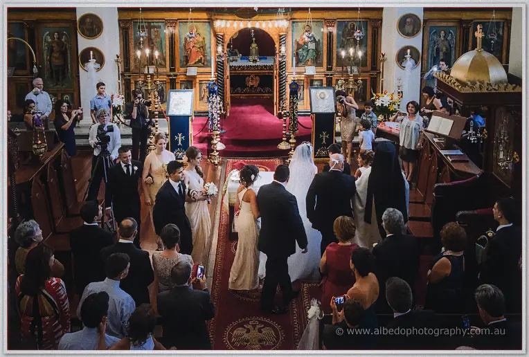 Elevated view of the crowning rite amid candlelit icons as photographers and guests crowd round