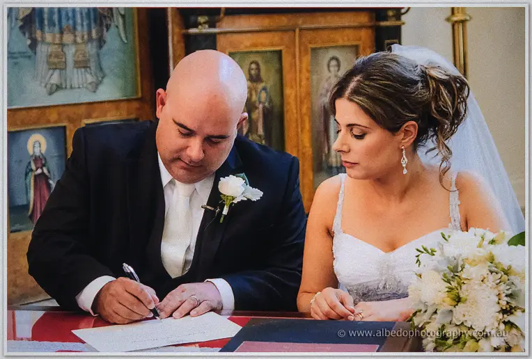 James signs the register as veiled Elena leans in among the timber icon screens