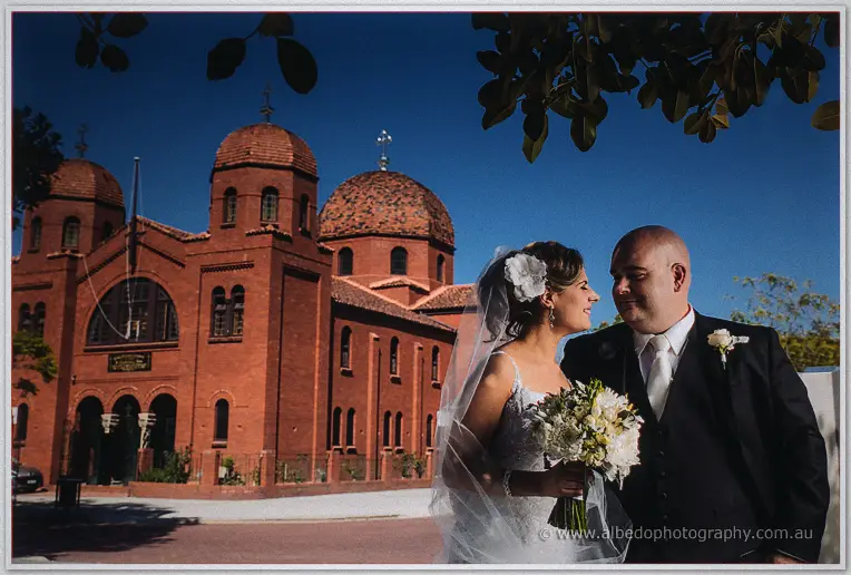 Elena gazes up at James as the domed red-brick church glows warmly behind them under a deep blue sky