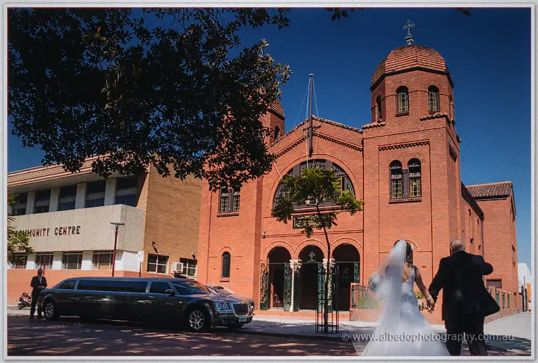 A bride in white and a man in a dark suit walk hand in hand toward the arched church entrance past a limousine