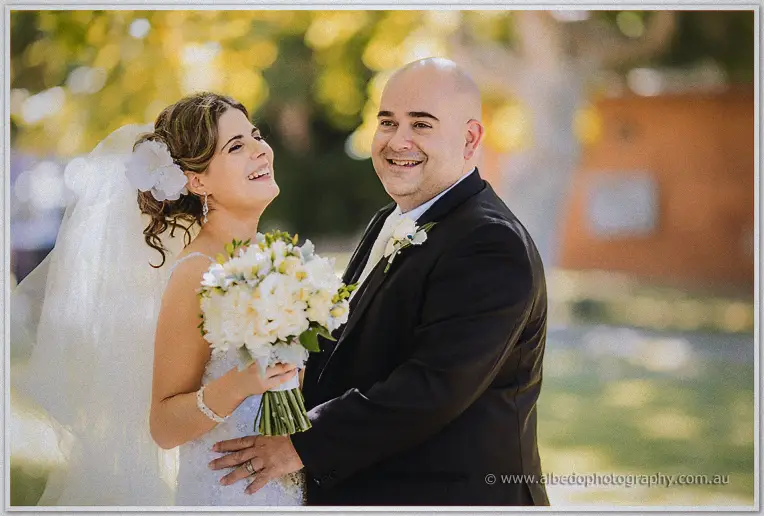 Elena laughs up at James while he holds her waist, her white peony bouquet glowing in soft parkland light