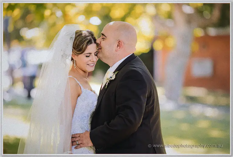 James kisses Elena's temple as she smiles softly, autumn foliage blurred gold behind them in the park