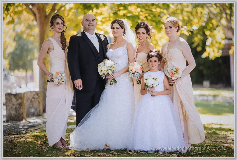 Elena, James, three bridesmaids in blush gowns and a flower girl gather beneath golden autumn trees