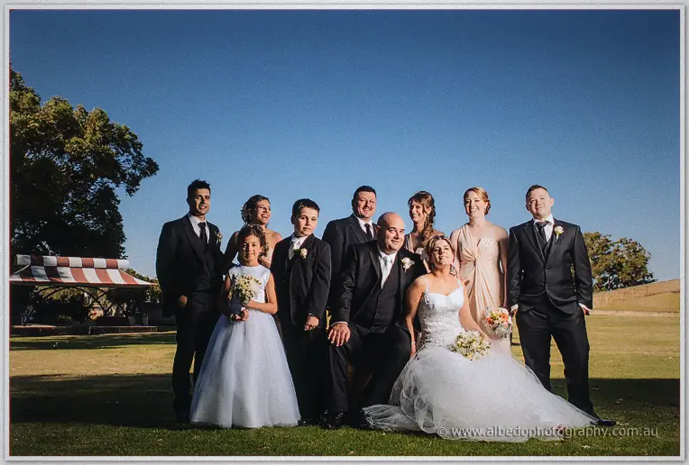 The full wedding party poses on a sunlit lawn, James and Elena seated at the front beneath a clear blue sky