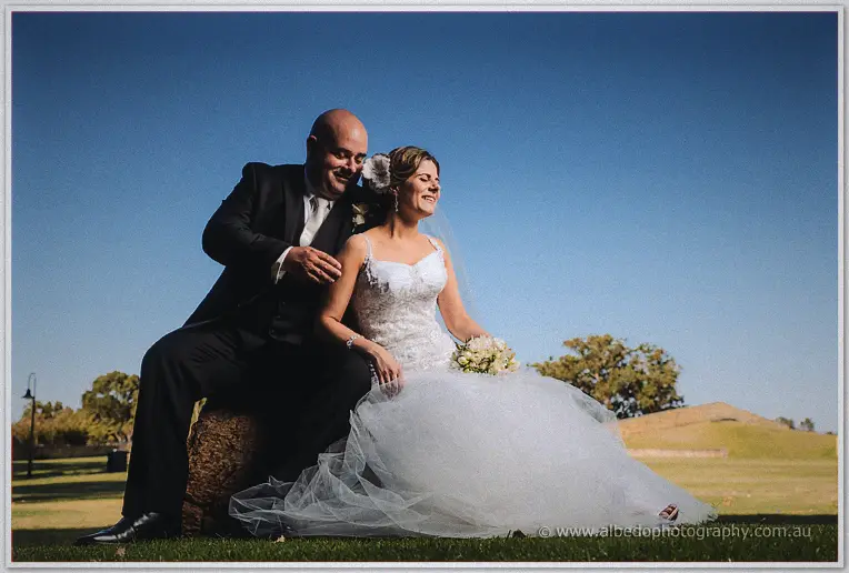 James and Elena sit laughing on a rocky outcrop, her tulle gown spread across the grass under a vast blue sky