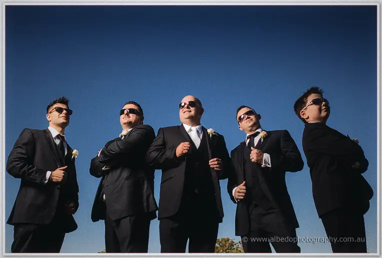 Shot from low down, James and four groomsmen in sunglasses strike cool poses against an empty blue sky