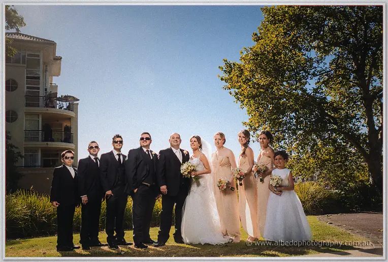 The whole bridal party lines up on a riverside lawn near Matilda Bay, apartment towers rising behind the trees