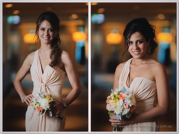 Two bridesmaids in pale peach halter gowns smile with peach and cream bouquets in a warmly lit hotel lobby