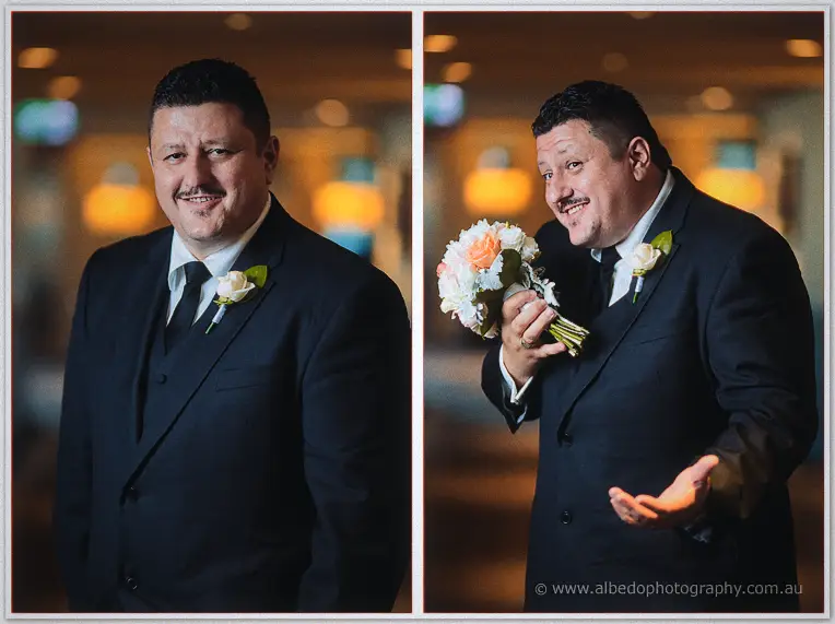 A grinning groomsman in a black suit smells the bridal bouquet and shrugs playfully in golden lobby light