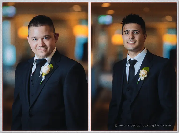 Portrait pair of two groomsmen in navy suits with white rose boutonnieres against softly blurred lounge lights