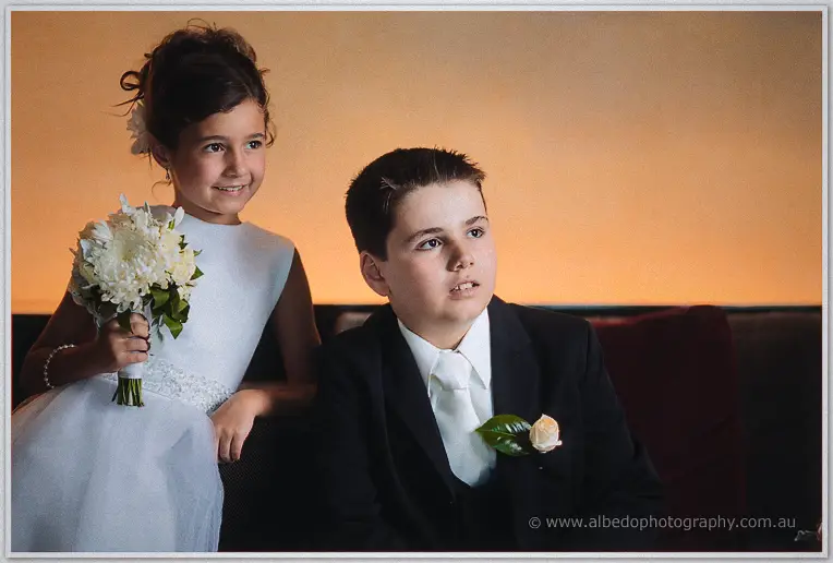 A flower girl clutching white blooms and a page boy in a suit gaze off camera against a warm amber wall