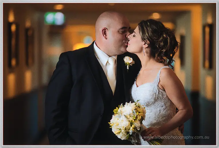 Elena kisses James's cheek in a softly lit hotel corridor, her bouquet of cream blooms in front of them
