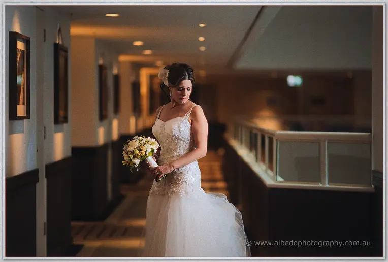 Elena gathers her lace gown along a dim hotel walkway, framed prints glowing in lamplight behind her