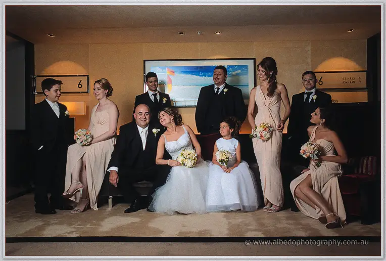 The wedding party gathers on sofas under a beach artwork, Elena and James seated centre with the flower girl