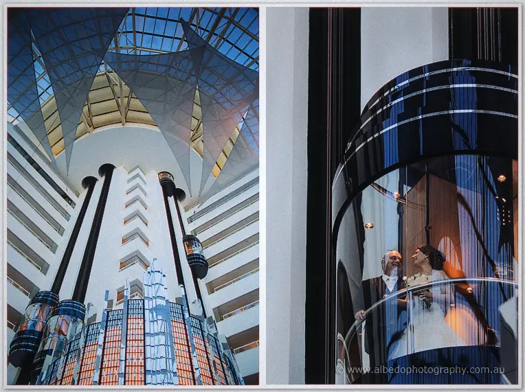 The couple rides a glass elevator through a soaring hotel atrium, balconies and sail-like canopy above them