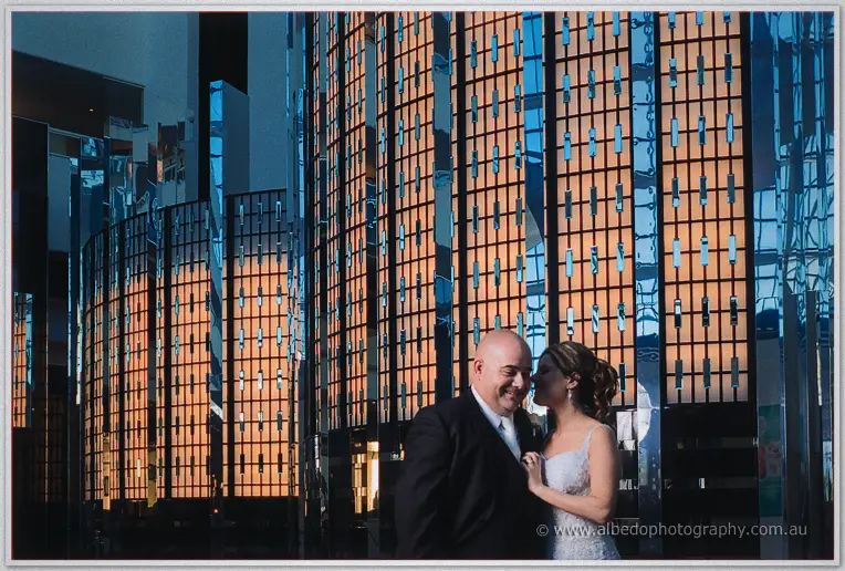 James and Elena smile close together against a striking wall of amber-lit mirror panels at dusk