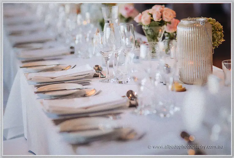 Crystal glasses and folded napkins line a white runner past pink roses and a ribbed silver lantern