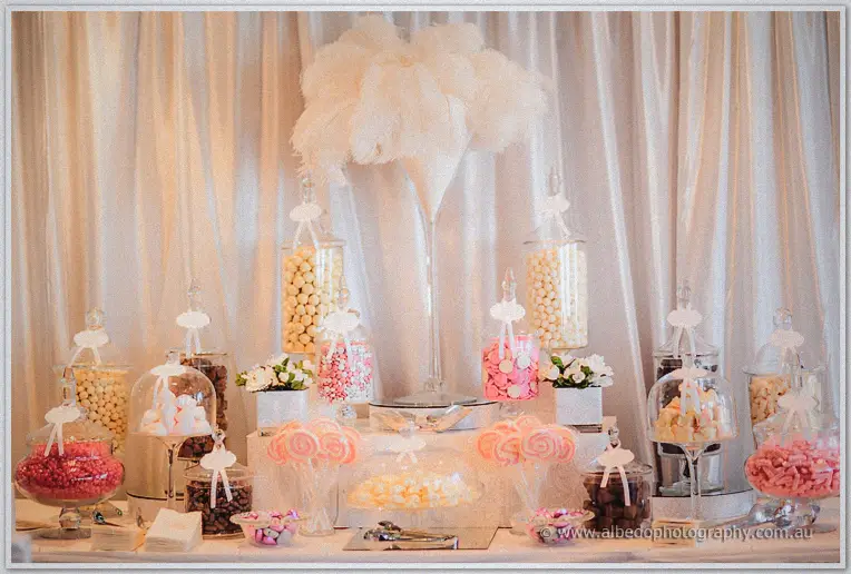 Glass jars of pink and cream sweets crowd a lolly buffet beneath towering white feathers on the dessert table
