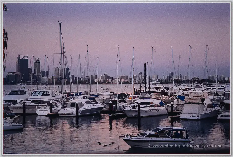 Yachts rest on glassy water at Matilda Bay as dusk turns the Perth skyline a soft violet