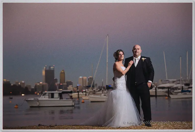 Elena straightens James's lapel on the shoreline as city towers and yacht masts glow behind them at dusk