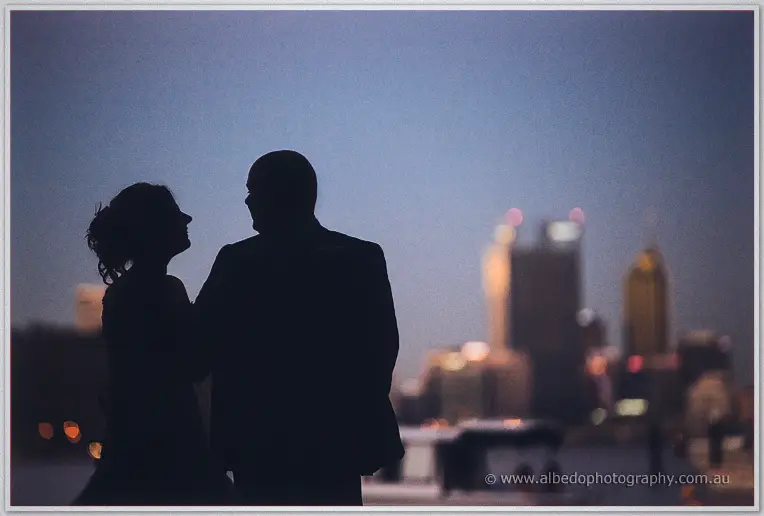 Elena and James stand silhouetted against the twilight sky, Perth's city lights blurred across the water