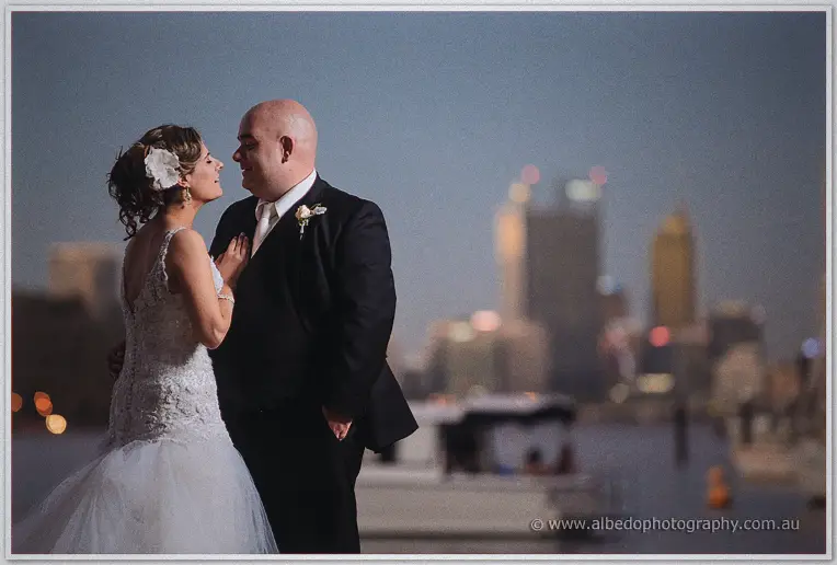 Elena touches James's chest as they laugh by the water, Perth's evening skyline a soft blur behind