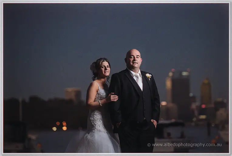 Elena holds James's arm by the marina at night, the softly lit city skyline floating behind them