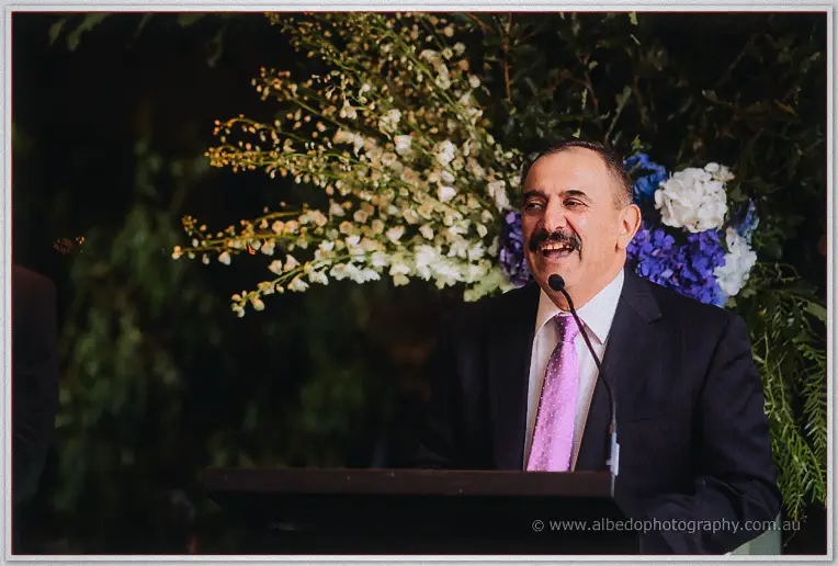 A moustached man in a pink tie laughs into the microphone beneath cascading blue and white flowers