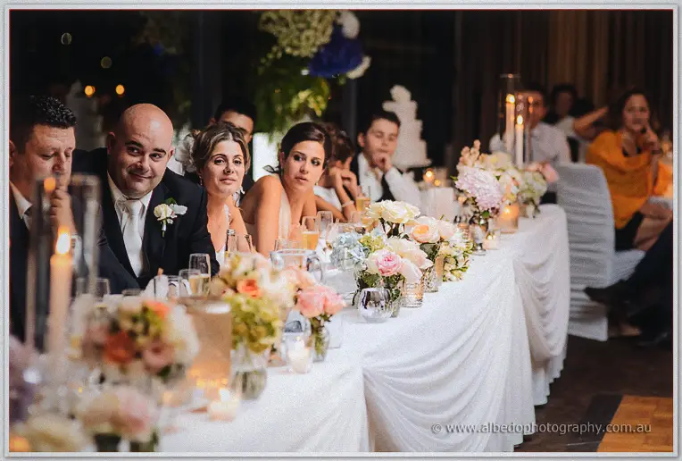 James grins at the camera along the bridal table while guests watch speeches under candlelight and blooms