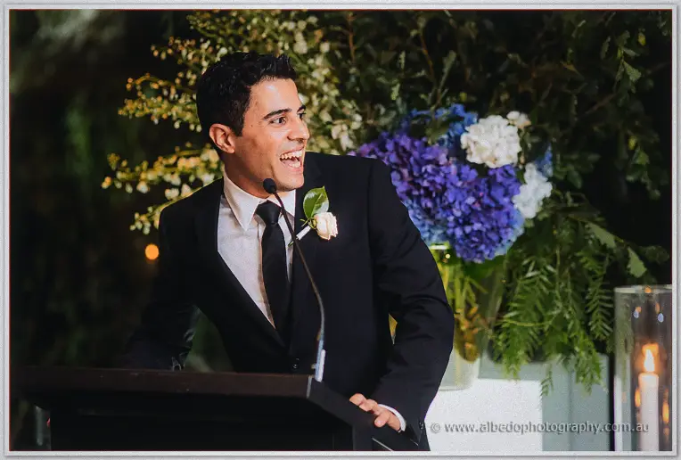 A groomsman in a black tie delivers an animated speech at the lectern in front of blue hydrangeas