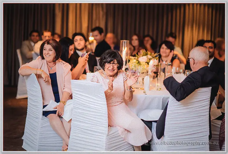 Wedding guests in blush and navy laugh together at candlelit round tables during the reception speeches
