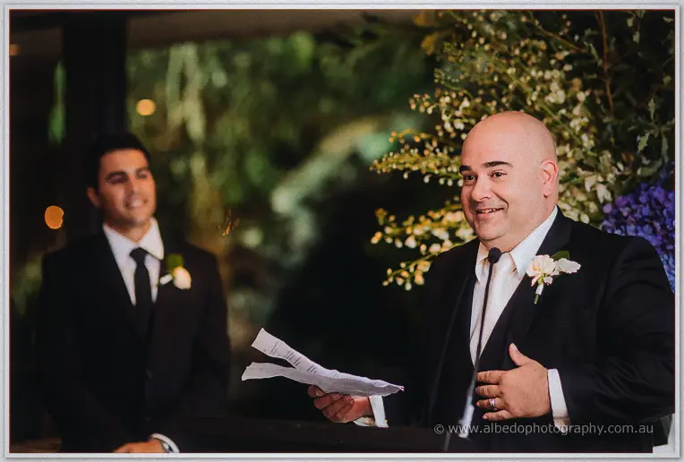 The best man wipes his eye mid-speech as a grinning groomsman watches under soft garden lights