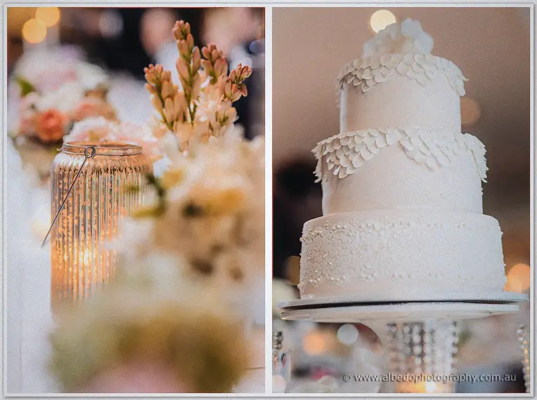 Ribbed candle lantern glowing beside peach blooms and a three-tier white cake dressed with sugar petals