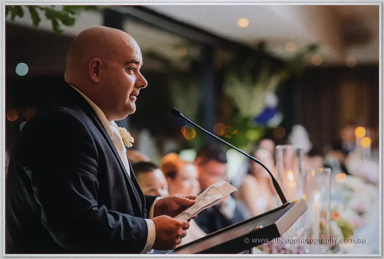 Groomsman with cream rose boutonniere reads his speech at a lectern in the candlelit Matilda Bay dining room