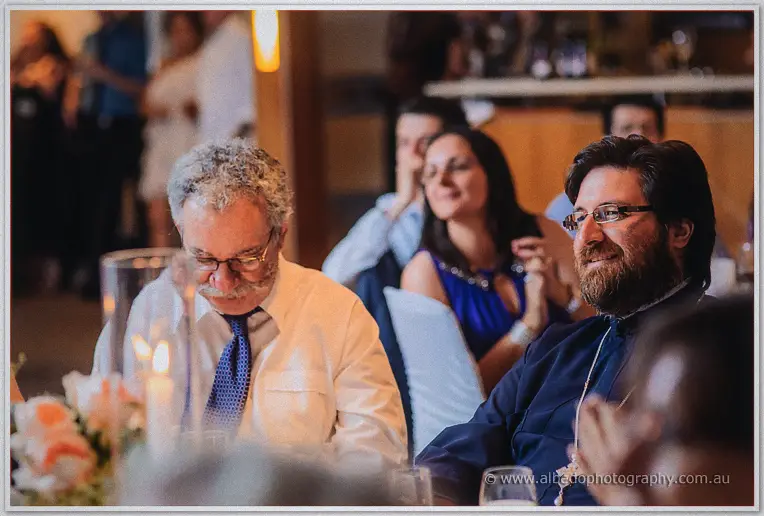 Wedding guests listening intently as a bearded priest in dark robes smiles during the Matilda Bay reception