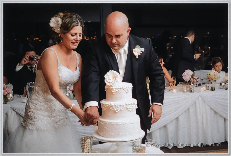 Bride with a white flower in her hair and groom in a black suit cutting their petal-tiered cake at Matilda Bay