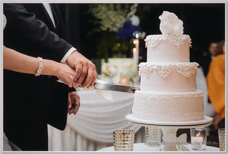 Close-up of the couple's joined hands guiding the knife through their blush-pink three-tier wedding cake