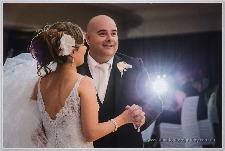 Groom in black suit and tie smiling at his bride in a beaded lace gown during their first dance
