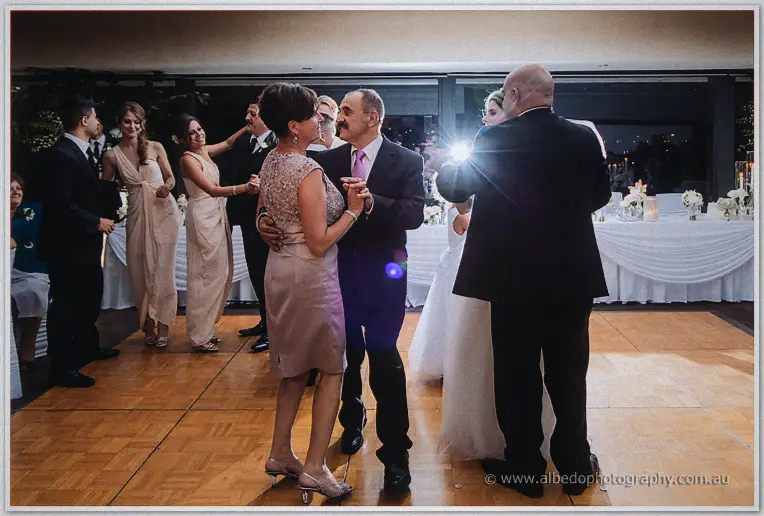 Parents dancing among guests in blush gowns on the wooden dance floor, camera flash flaring behind them
